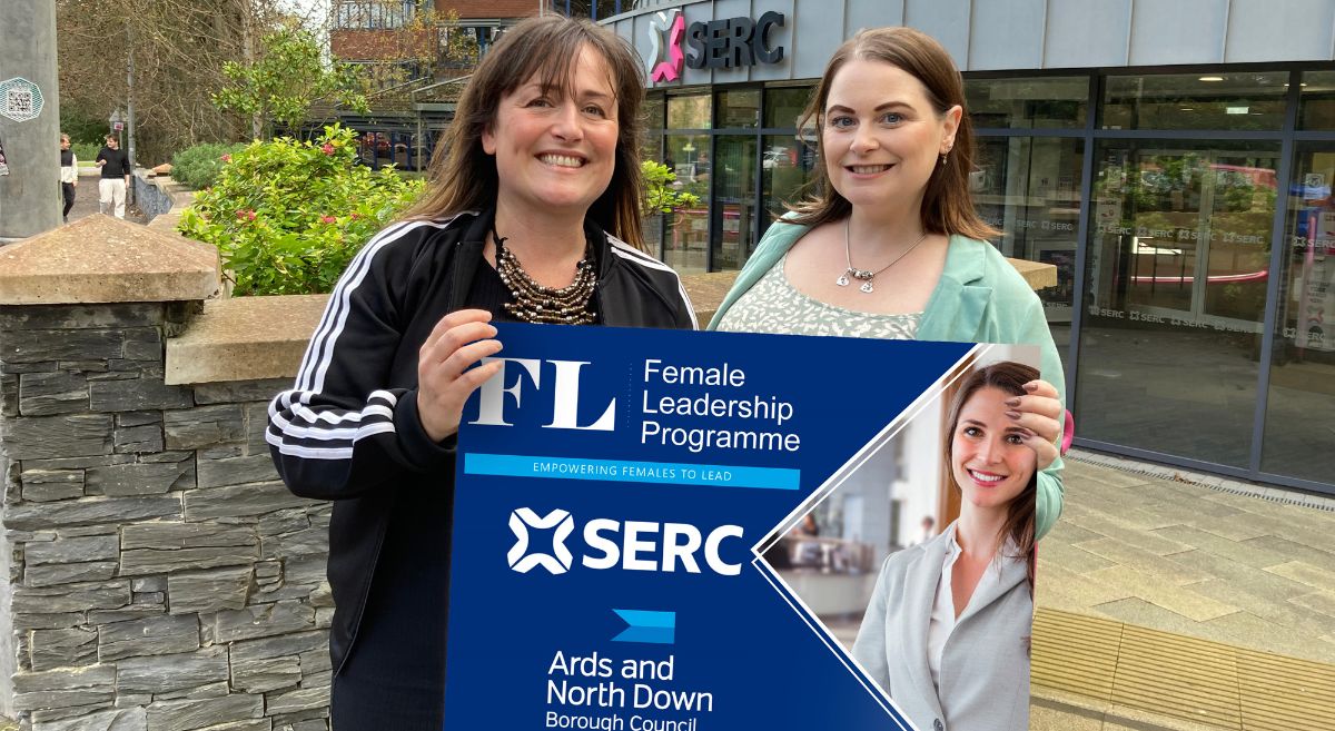 Two women, with dark hair,  outside SERC's Bangor Campus holding  a sign that says Female Leadership Programme, shows a female in office attire with SERC and Ards North Down Council logos